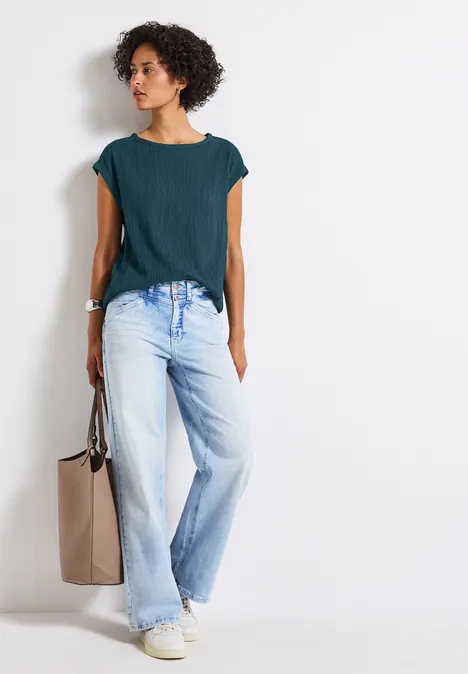 Frau mit lockigen Haaren trägt grünes Oberteil, weite Jeans und weiße Sneaker, hält eine beige Tasche.